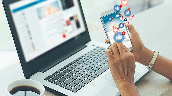Computador en el fondo y una persona navegando en su celular en las redes sociales